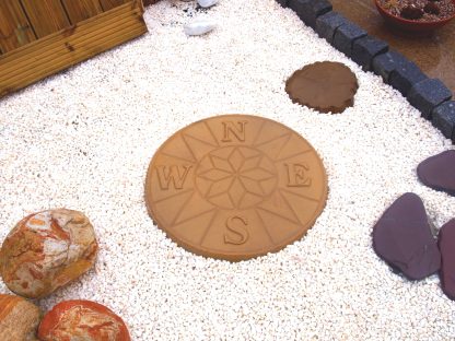 buff gold compass stepping stone on a white gravel path. there are red lake rocks to the left and purple slate rockery to the right of the image.